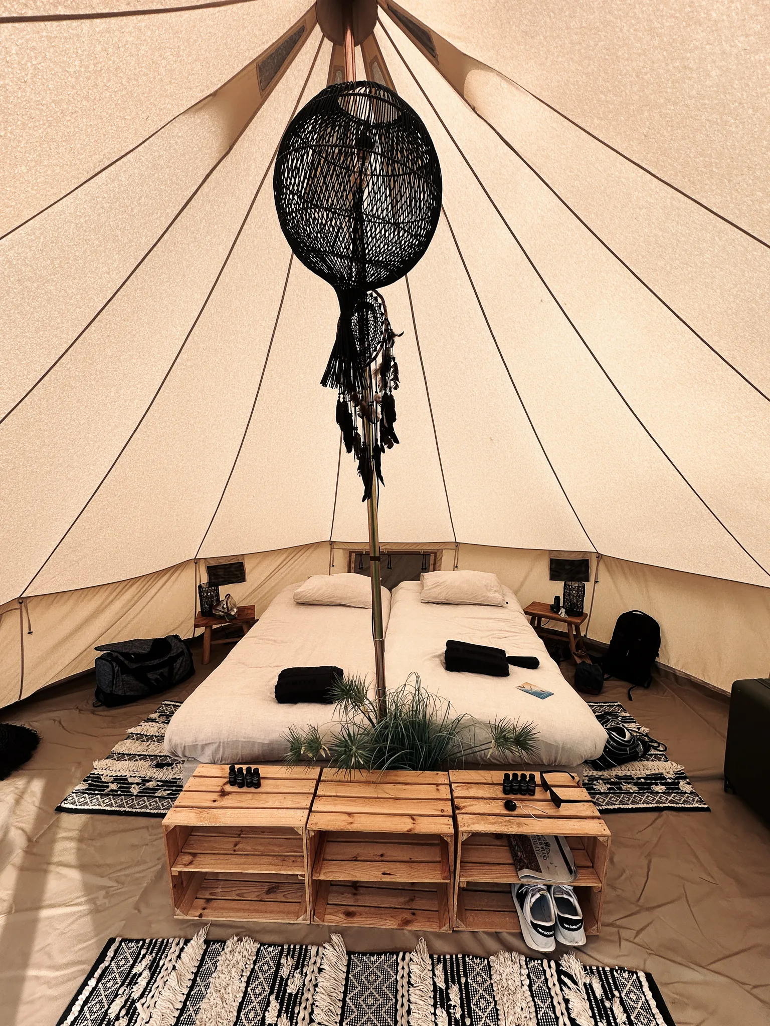 Inside a bell tent - two mattresses pushed together under a black wicker lamp hung from the centre pole, folded towels, wooden crates for shelves, patterned rugs over the groundsheet.