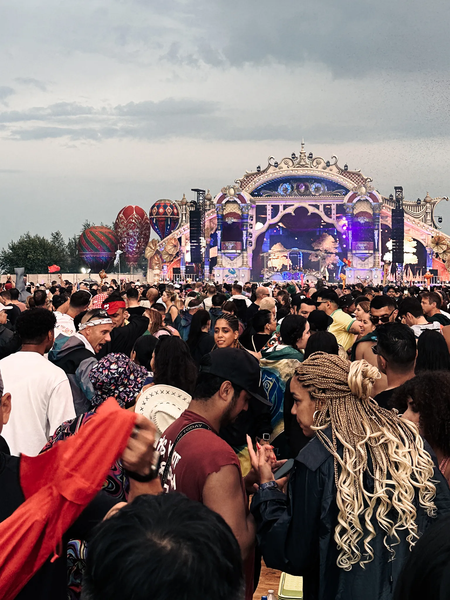 A stage with a gilded arched facade lit purple under heavy grey evening cloud, hot-air balloons tethered off to the left, a dense crowd filling the field in front of it.