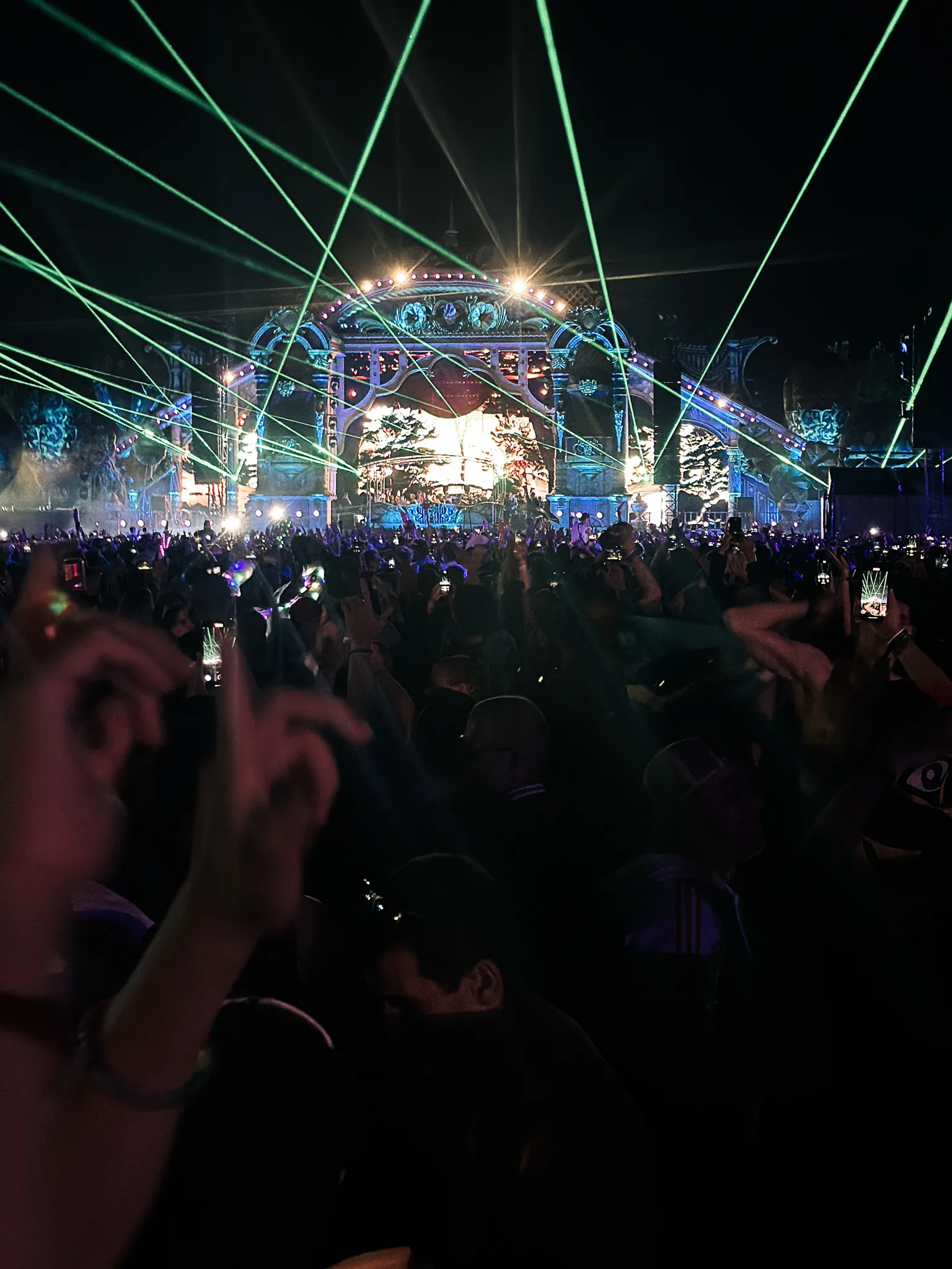 The same arched stage at night, green lasers fanning out over the crowd from the arch, phones held up everywhere, a hand in the foreground making a sign against the light.