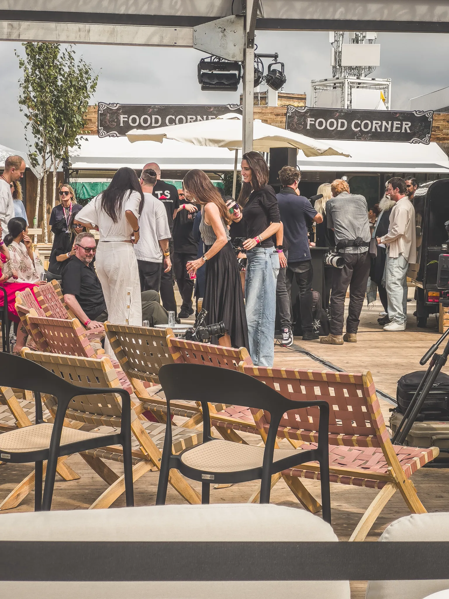 A shaded seating area of wooden deck chairs under two Food Corner signs, people standing around chatting mid-afternoon, a photographer with a camera slung at his hip.
