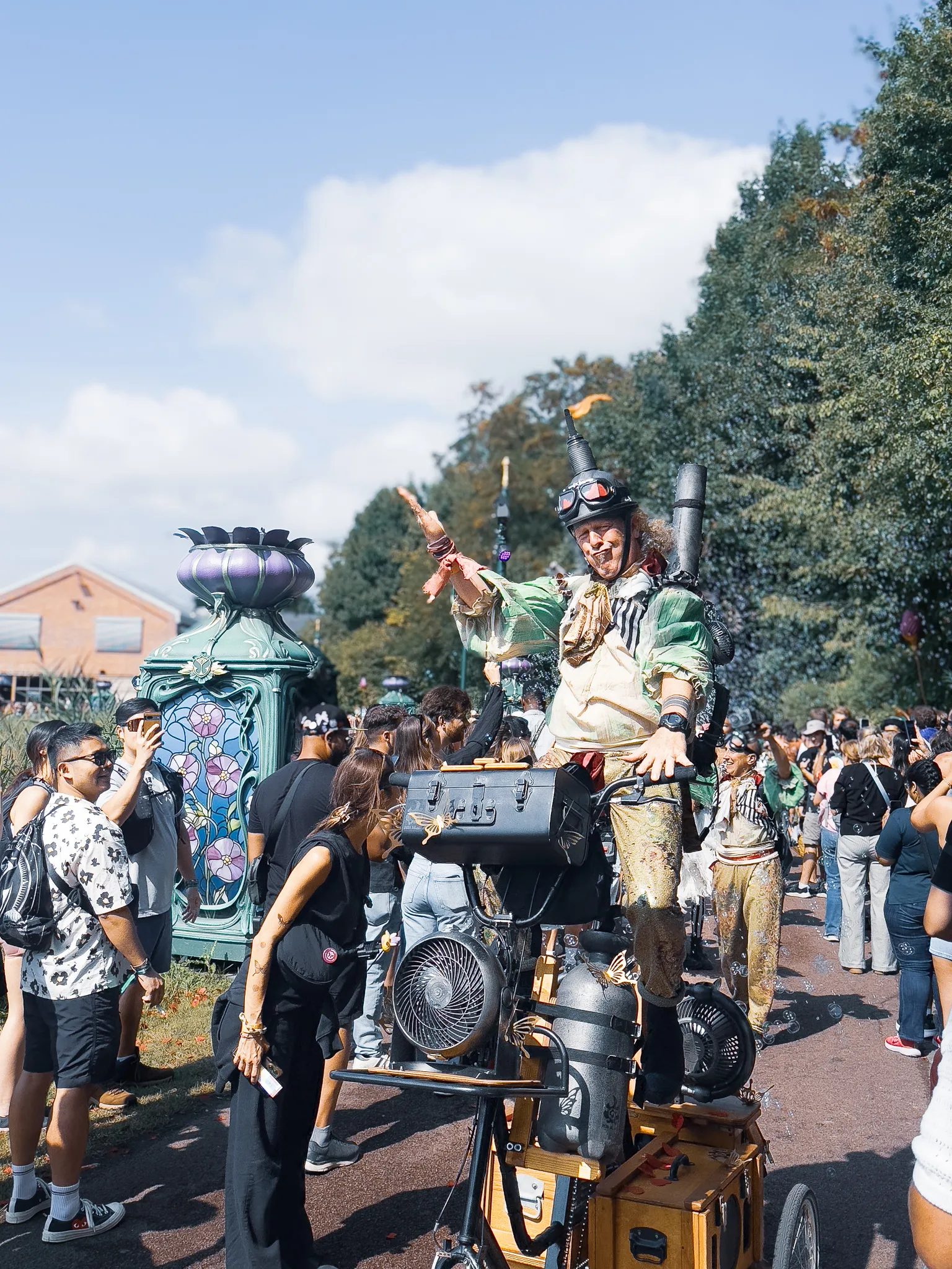 A street performer in goggles and a patched green costume riding a homemade contraption of fans, tanks and suitcases, waving to the crowd on the path, an art nouveau lamp behind.