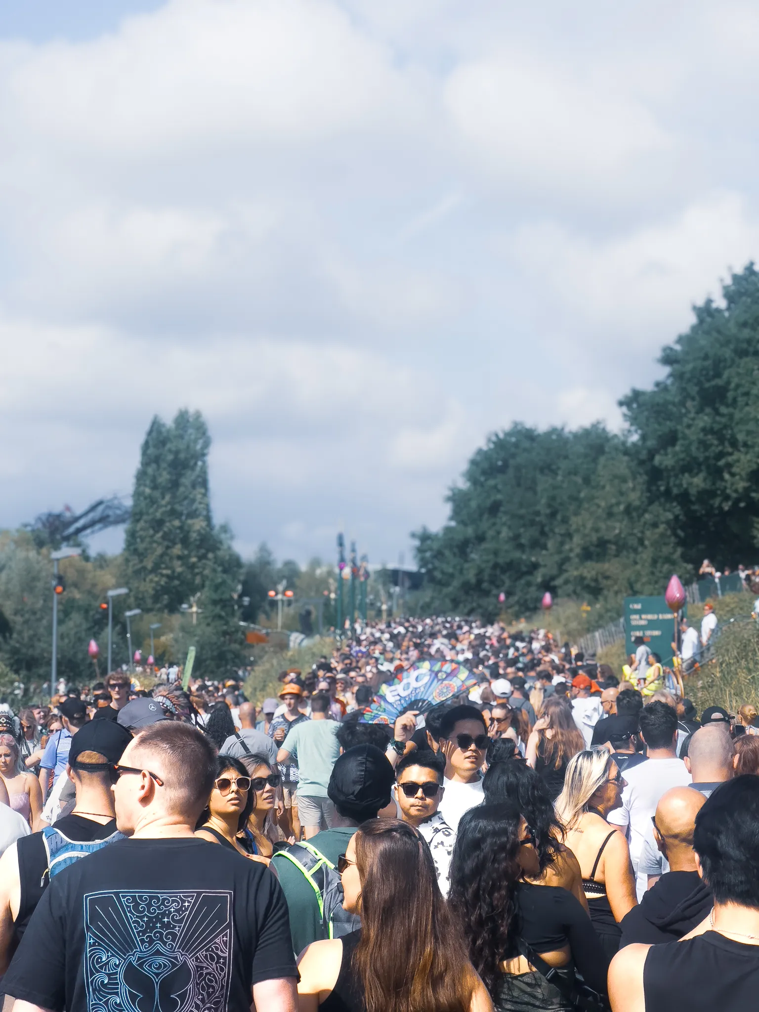 The path between stages packed shoulder to shoulder under a white sky, a painted fan raised above the heads, trees and a crane on the horizon.