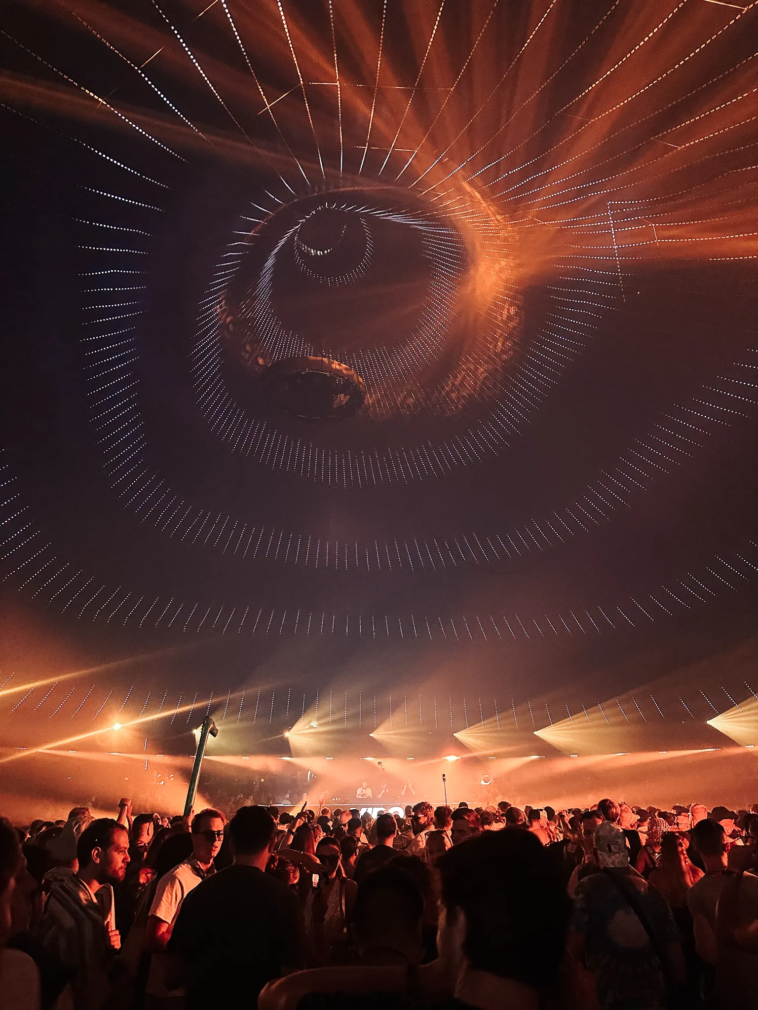 Inside a tent stage, a huge eye picked out in strings of lights across the canopy, amber haze below it, the crowd facing a booth at the far end.