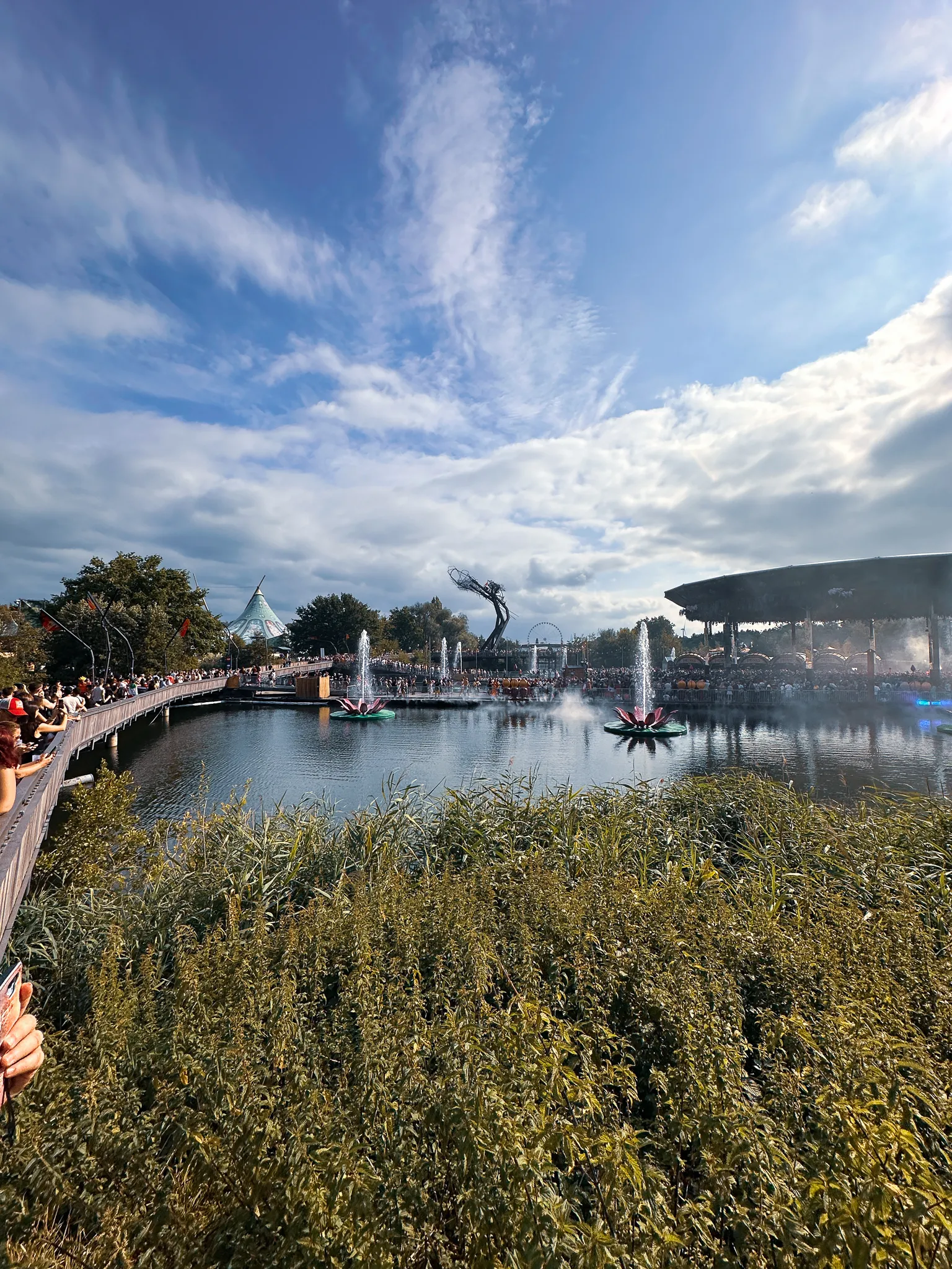 The lake in afternoon sun - fountains playing between lotus-shaped floats, a boardwalk of people along the left bank, a giant statue and a wide stage roof across the water.