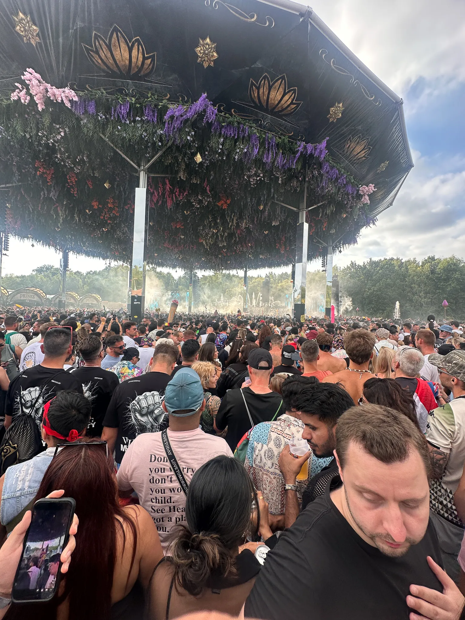Under a stage canopy hung with purple and pink blossom and gold lotus emblems, the crowd shoulder to shoulder beneath it, someone in front holding up a phone to the view.