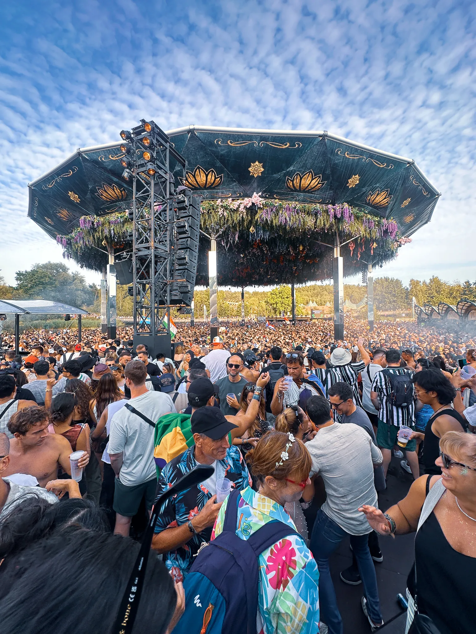The blossom canopy from further back under a mackerel sky, the crowd running all the way to the trees, an Indian flag up on a pole, a man in a floral shirt turning to the camera.