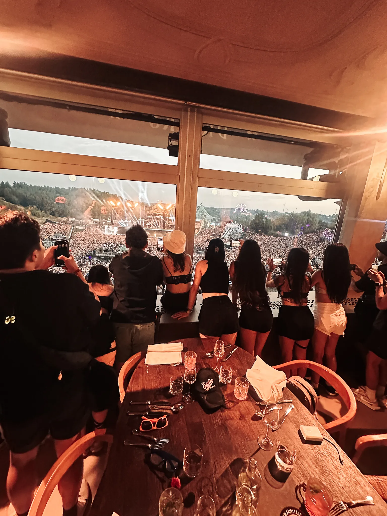 From a table by a window high above the grounds - a row of people at the glass looking down on a stage lit up across the field at dusk, wine glasses and cutlery still on the table.