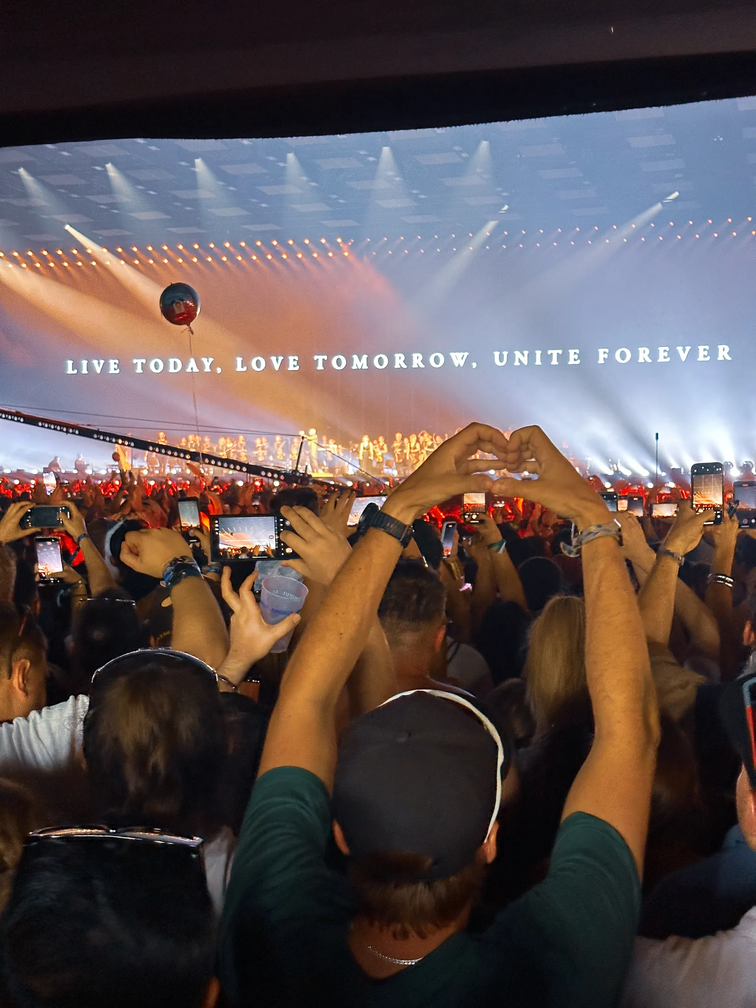 Inside a stage, the screen reading Live Today, Love Tomorrow, Unite Forever above rows of performers and a balloon, a pair of hands making a heart in the foreground, phones everywhere.
