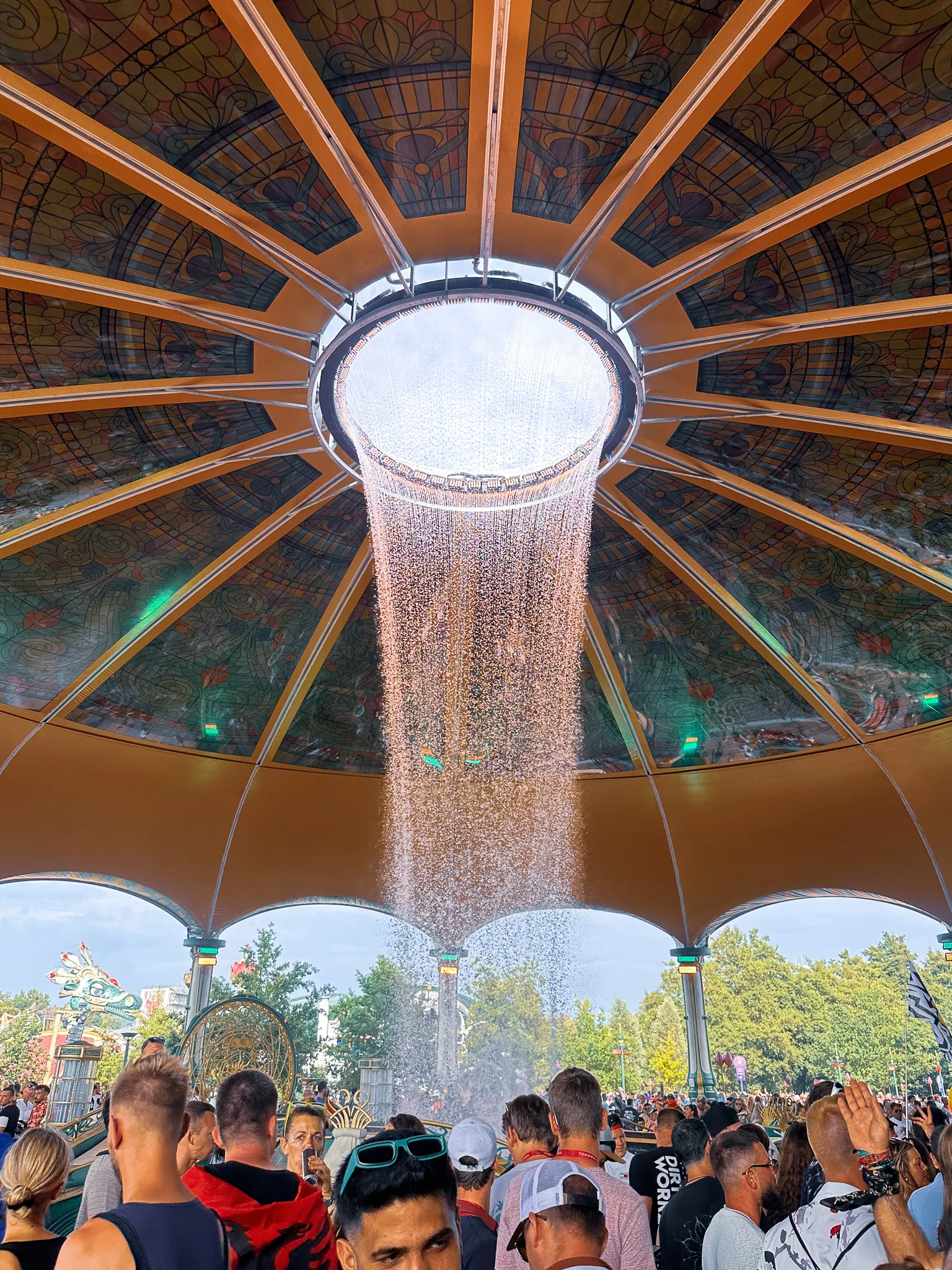 Under a stained-glass dome, a ring of water falling from an opening in the centre onto the crowd below, decorated pavilions and trees through the open sides.