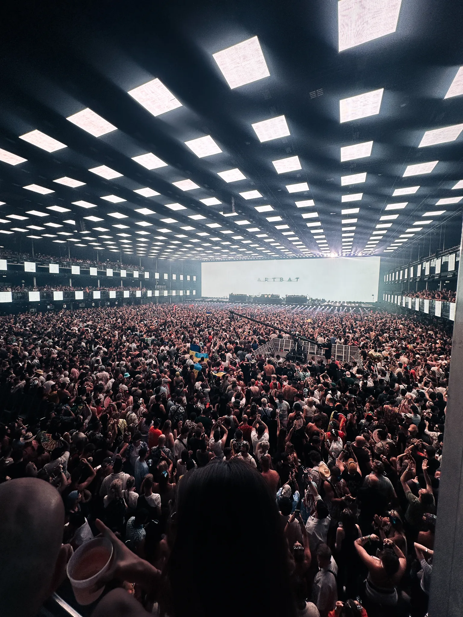 A vast indoor hall from the balcony, a grid of white light panels the whole length of the ceiling, the screen at the far end reading ARTBAT, thousands on the floor below.