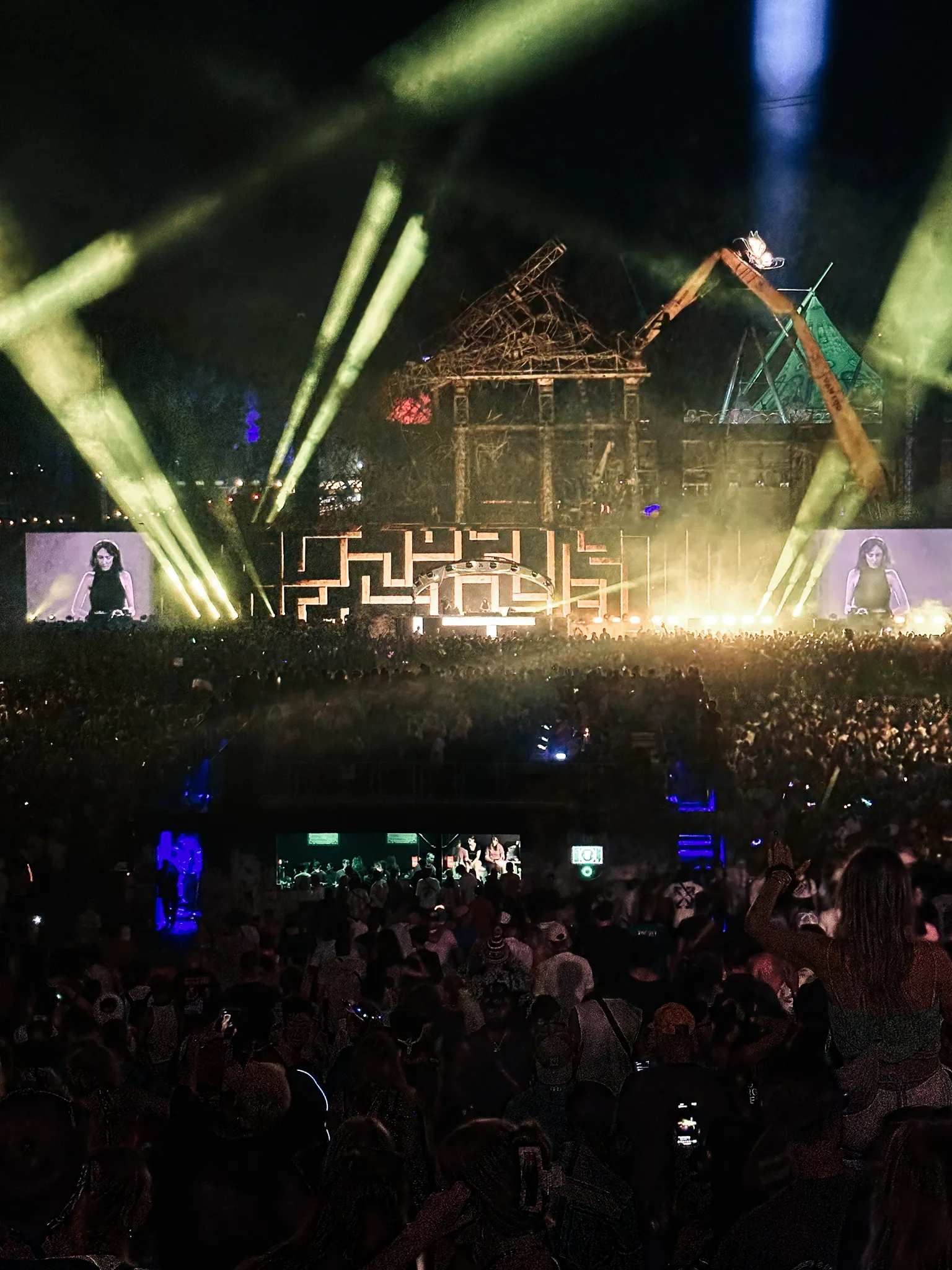 A night stage built of raw timber like a broken tower, yellow beams crossing above it, a maze of light around the booth, the DJ on screens either side, the crowd reaching back into the dark.