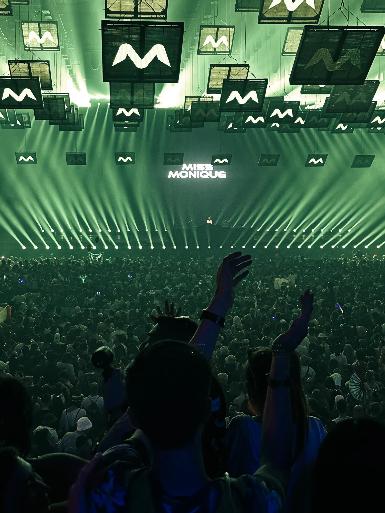 An indoor stage lit green, the screen reading Miss Monique, a grid of hanging boxes with a wave logo overhead, fans of light beams over the floor, hands up in front.
