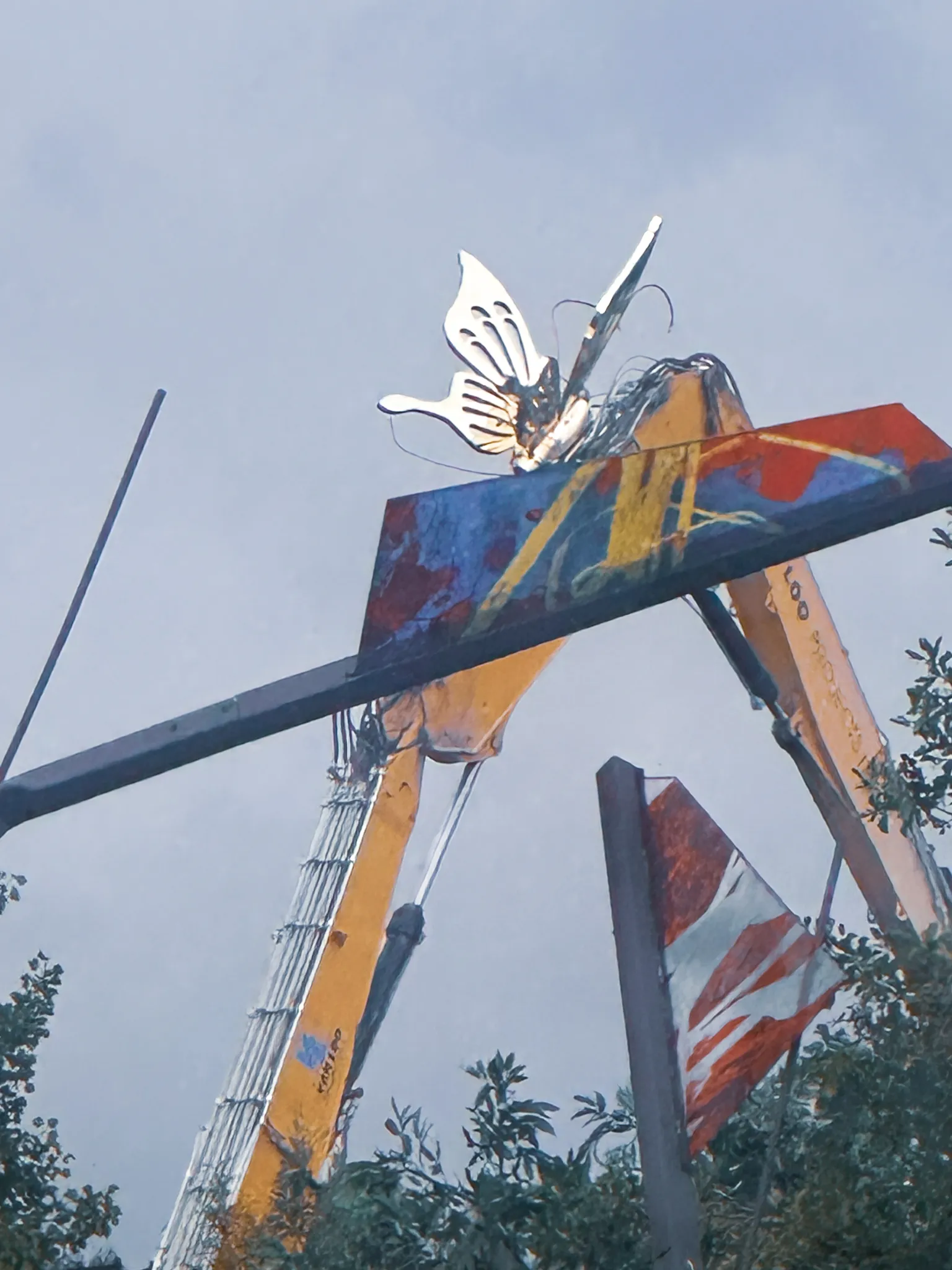 A cut-out butterfly perched on the boom of a crane, painted panels hanging off it, a striped flag and treetops against a grey evening sky.