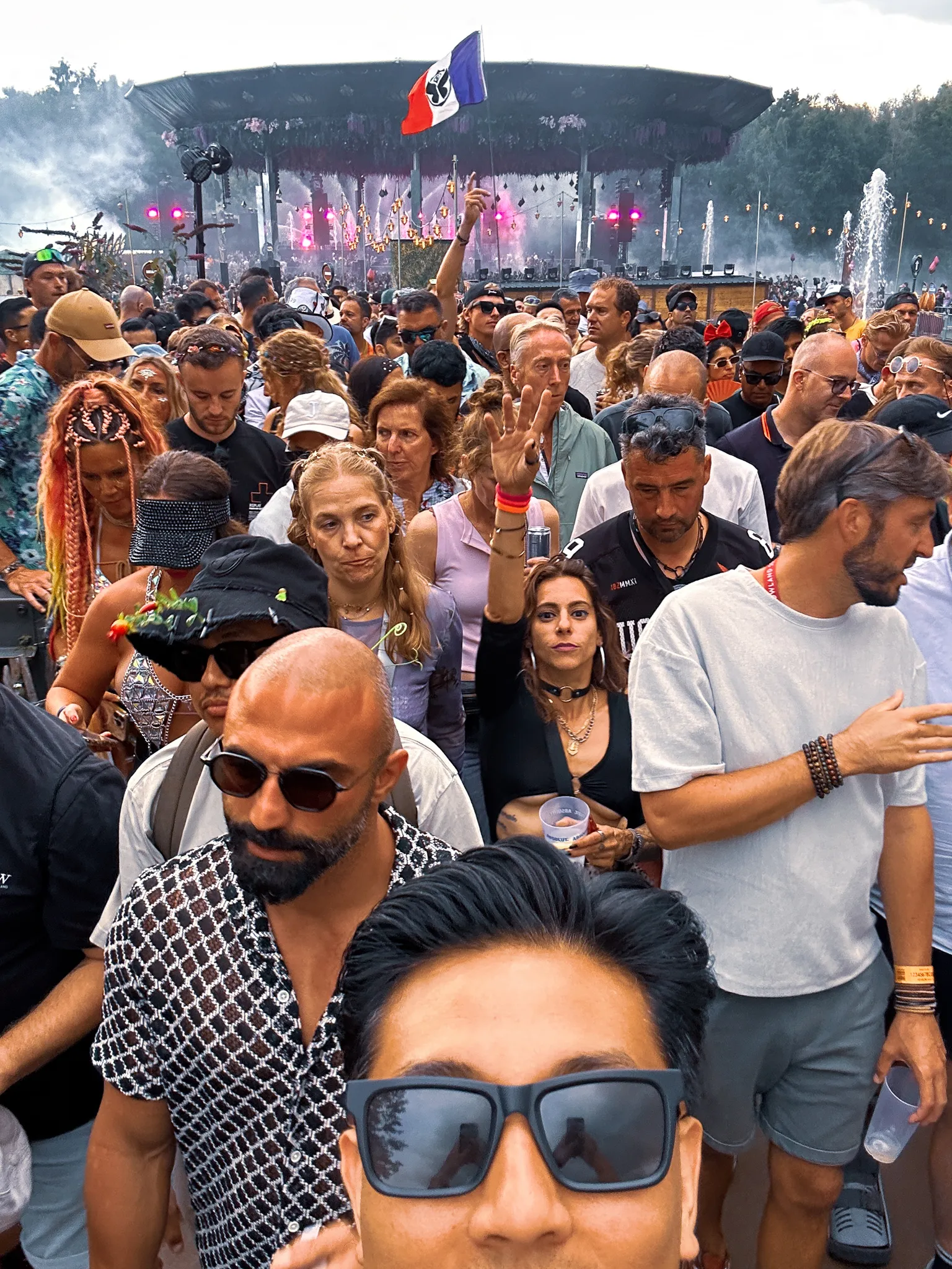 A selfie from inside the crowd - sunglasses in the foreground, a man in a mesh shirt and a woman holding a drink behind, a French flag up on a pole, the blossom canopy at the far end.