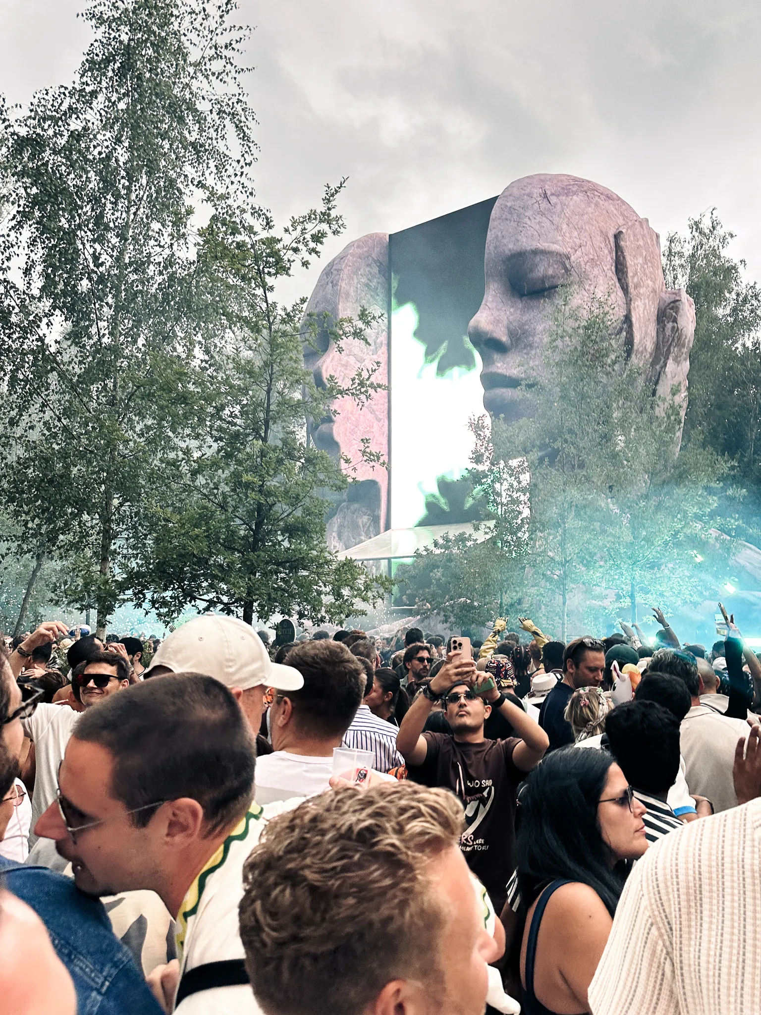 Two giant sculpted faces in profile with a screen between them, blue smoke drifting across the crowd below, birch trees at the left, the last light going grey.